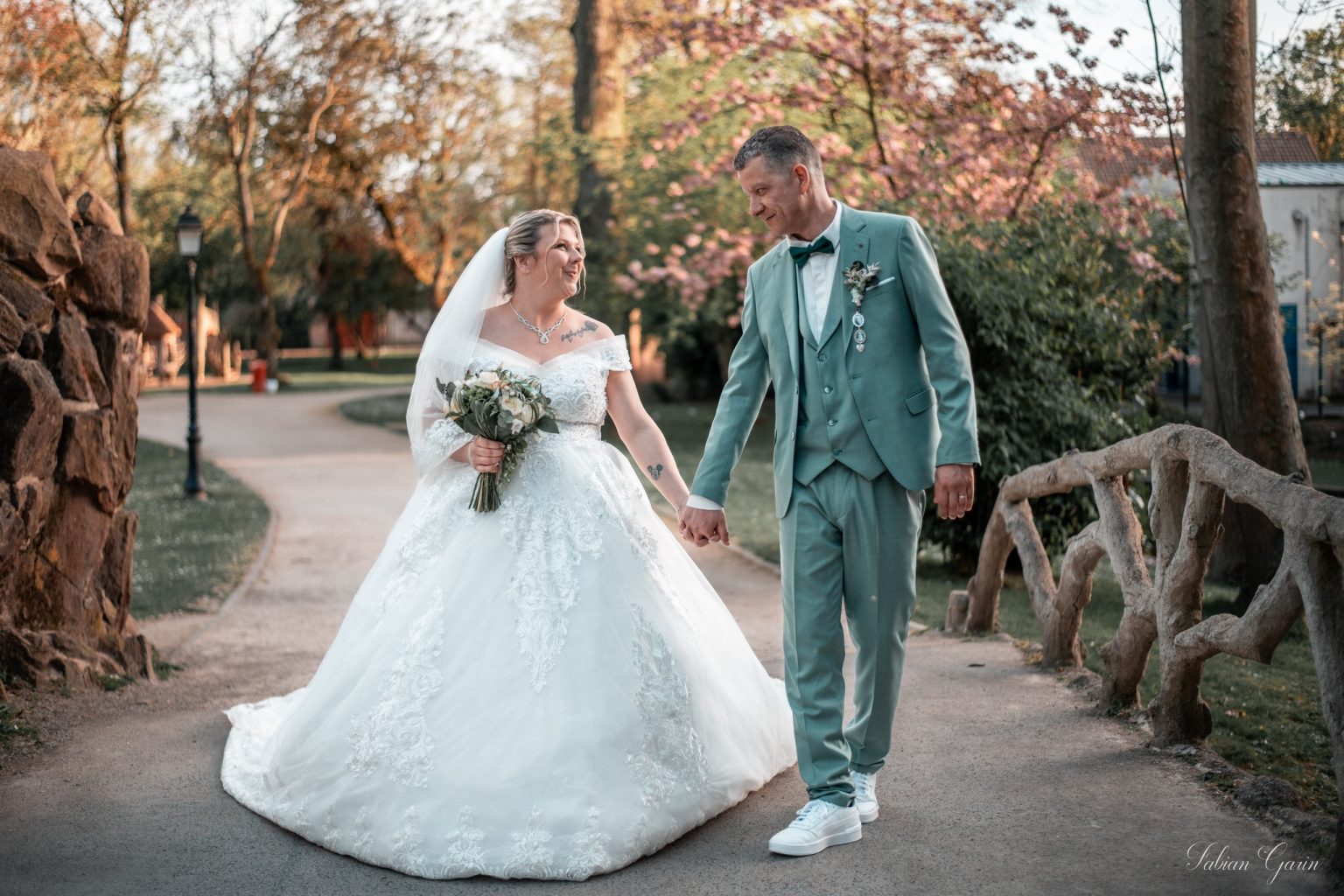 Julianna et Meadi marchent main dans la main dans un parc fleuri, en tenue de mariage, se regardant avec tendresse après leur cérémonie.