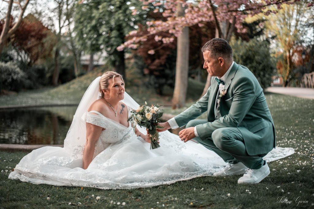 Le marié tend un bouquet à la mariée assise dans l’herbe, tous deux en tenue de cérémonie, entourés de verdure et de fleurs, lors de leur mariage au printemps 2024.
