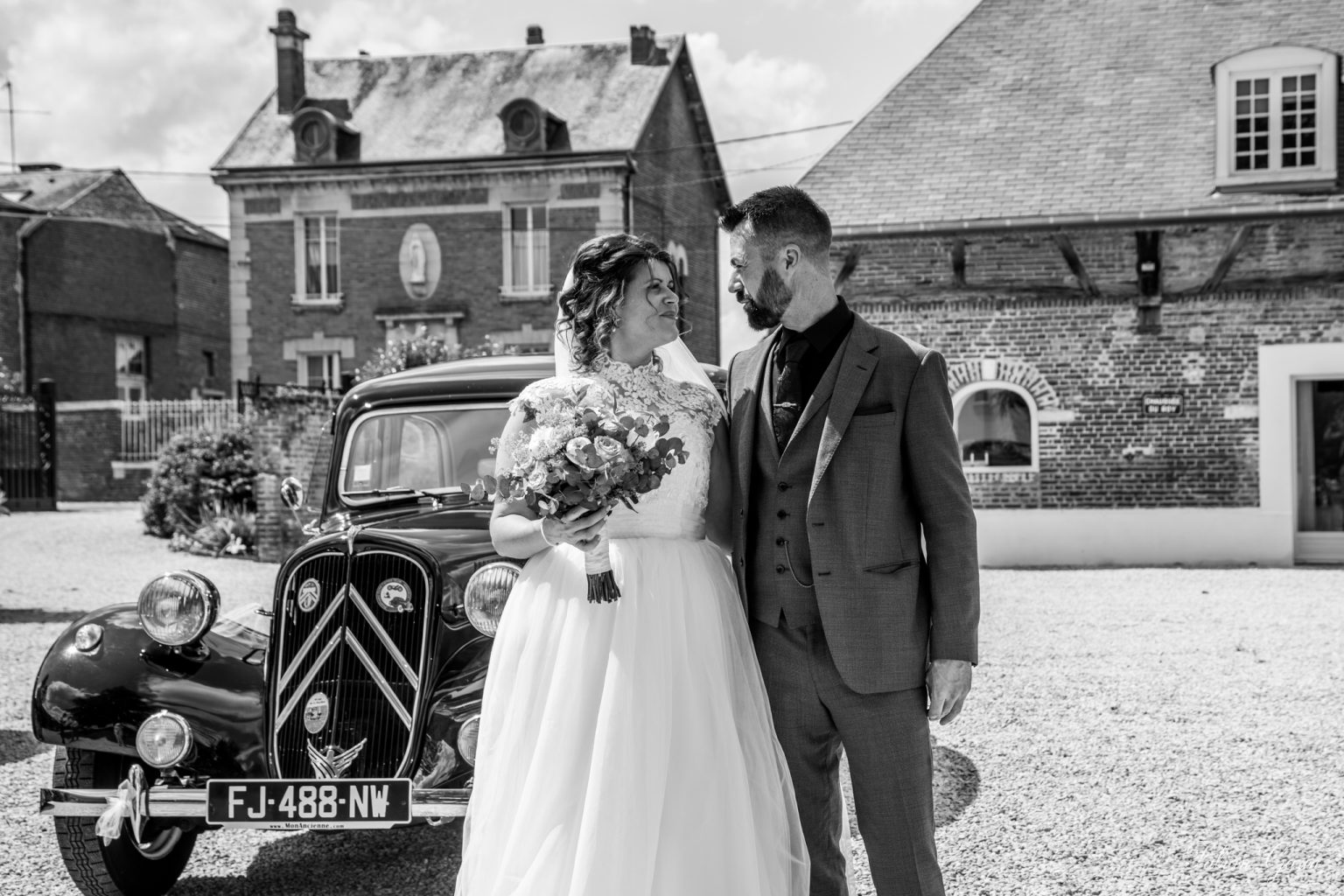 Charlotte et Pierre se regardent tendrement devant une voiture de collection noire, le jour de leur mariage, à la mairie de Beaurains-lès-Noyon.