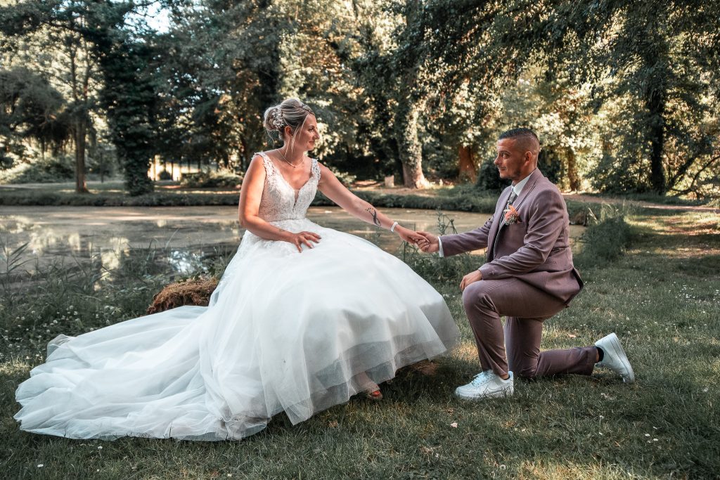 Jennifer, assise en robe de mariée devant un étang, tend la main à Christopher, en costume parme, agenouillé face à elle, dans un décor verdoyant à Vendin-le-Vieil.