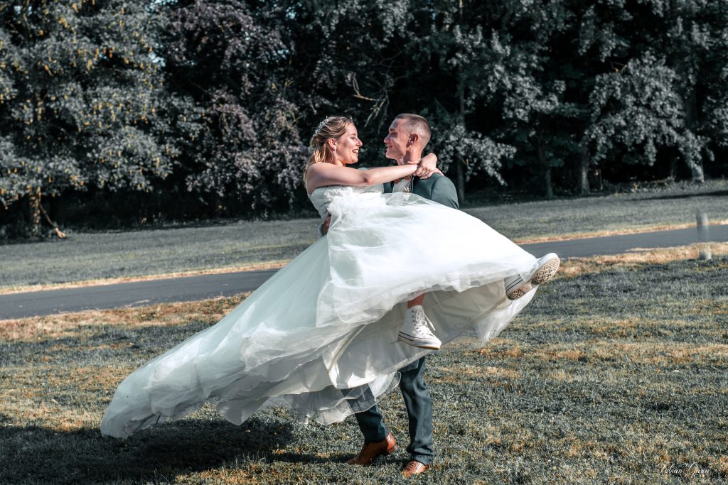 Le marié porte sa femme dans ses bras avec tendresse dans un parc, devant les arbres, au Château de la Motte Fénelon à Cambrai. La mariée rit en le regardant, en robe blanche et baskets, dans une ambiance joyeuse et naturelle.