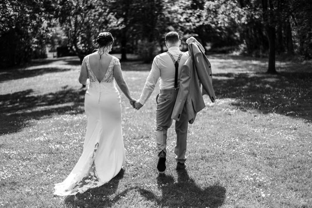 Les mariés, de dos, marchent main dans la main à travers un parc ensoleillé, dans une atmosphère paisible et romantique, capturée en noir et blanc.